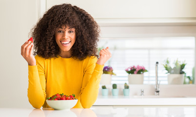 African american woman eating strawberries at home screaming proud and celebrating victory and success very excited, cheering emotion