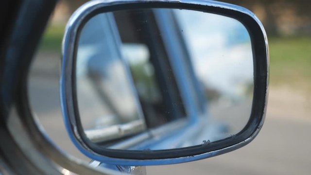 View to side mirror of old retro car with blurred city at background. Vintage automobile driving at the city street on sunny summer day. POV Close up Slow motion