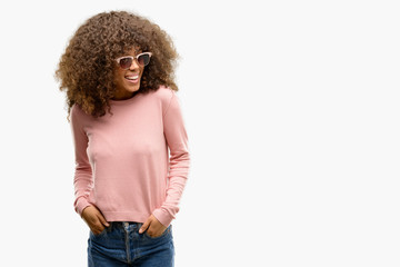 African american woman wearing pink sunglasses looking away to side with smile on face, natural expression. Laughing confident.