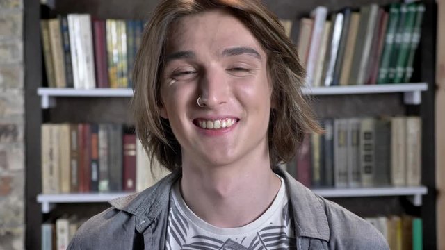 Young long hair man with nose ring is watching at camera, giggling, library on background