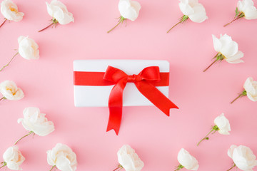 Gift box with red ribbon in the center on pastel pink background. Beautiful, fresh white roses. Giving or receiving a lovely gift. Celebration day. Different festive congratulation concept.