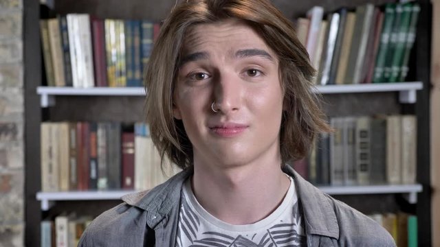 Young long hair man with nose ring is watching at camera, express acceptance, library on background