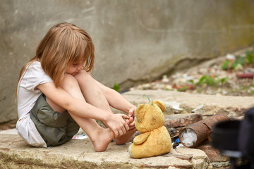 Orphan with bare feet in a shabby clothes playing with used teddy bear on the street.