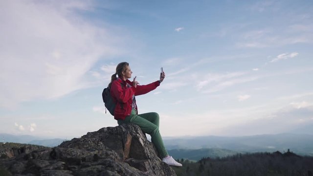 the traveler takes a selfie in the mountains. young female tourist sitting on the edge of a cliff and taking pictures on the camera smartphone