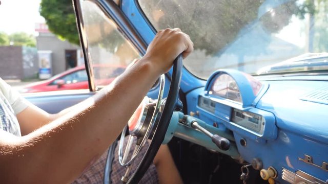 Unrecognizable man holding hands on a steering wheel and drives a retro car on the country road on sunny summer day. Young guy inside his old blue automobile. Close up Side view Slow motion