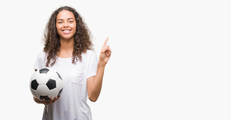 Young hispanic woman holding soccer football ball very happy pointing with hand and finger to the side