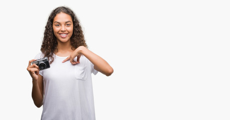 Young hispanic woman taking pictures using vintage camera with surprise face pointing finger to...