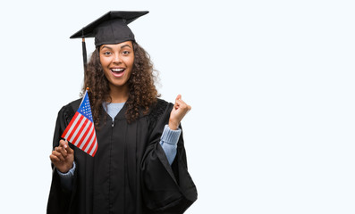 Young hispanic woman wearing graduation uniform holding flag of United States screaming proud and...