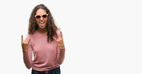 Beautiful young hispanic woman wearing sunglasses shouting with crazy expression doing rock symbol with hands up. Music star. Heavy concept.