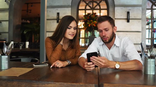 Stylish guy in eyeglasses and beautiful girl spending time together at cafe, watching a movie on smartphone in headhone, drinking coffee with dessert. Time with pleasure.