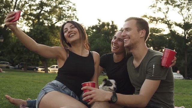 Portrait of three friends sitting on the grass in the park and posing for photo. Brunette girl taking picture with her smartphone. Friends together with small dog outdoors. Smiling, happy young people
