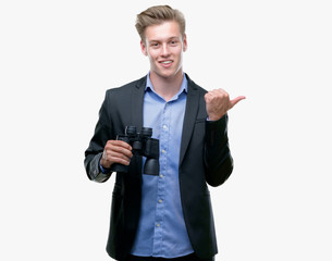 Young handsome blond man looking through binoculars pointing with hand and finger up with happy...