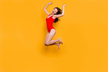 Fototapeta na wymiar Finally, summer! Full-body portrait of crazy brunett girl in red body with closed eyes jumping clenching her hands in fists and joyfully screaming isolated on yellow background