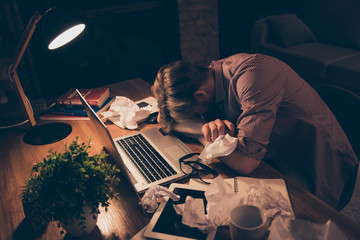 Portrait of exhausted, attractive, bored, tired man in shirt putting face on arms sitting at desk in work place, station having crumpled drafts on the table, working at night time - obrazy, fototapety, plakaty
