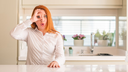 Redhead woman at kitchen doing ok gesture shocked with surprised face, eye looking through fingers. Unbelieving expression.