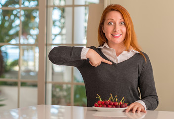 Redhead woman eating cherries at home with surprise face pointing finger to himself
