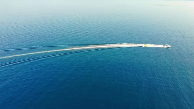 Aerial view of high speed boat pulls the banana boat on blue sea. Drone shot flying over people who enjoy in water sports