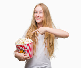 Blonde teenager woman eating pop corn very happy pointing with hand and finger