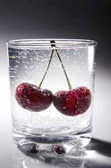 a glass of fresh, chilled mineral water from a mountain spring with two juicy, ripe, red cherry berries on a gray luminous background with reflection
