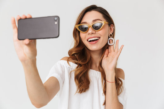 Photo of pretty european woman 20s wearing sunglasses taking selfie on smartphone, and touching earring, isolated over white background