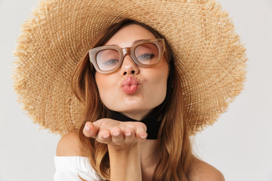 Portrait of adorable seductive woman 20s wearing big straw hat and sunglasses blowing air kiss at camera, isolated over white background