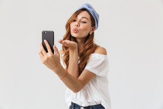 Photo of attractive joyous woman 20s smiling and blowing air kiss while taking selfie on mobile phone, isolated over white background