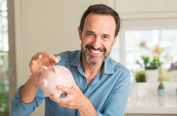 Middle age man save money on piggy bank with a happy face standing and smiling with a confident smile showing teeth - obrazy, fototapety, plakaty