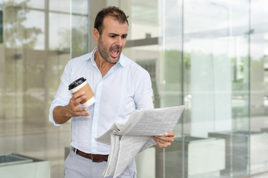 Furious business leader angry with negative review on his company. Mid adult Latin holding carton cup of coffee outdoors and yelling at newspaper. Bad news concept