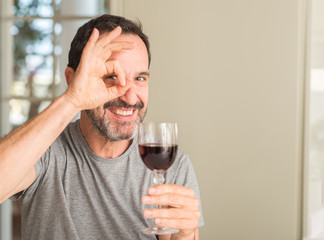 Middle age man drinking a glass of wine with happy face smiling doing ok sign with hand on eye looking through fingers