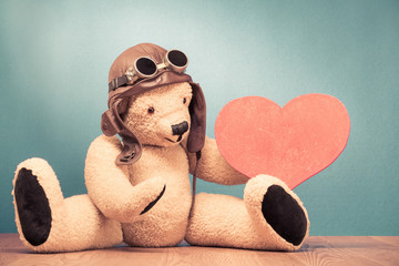 Retro Teddy Bear toy in leather pilot's hat and vintage goggles sitting on the floor with handmade wooden Valentine's day love heart shape front mint green wall background. Old style filtered photo