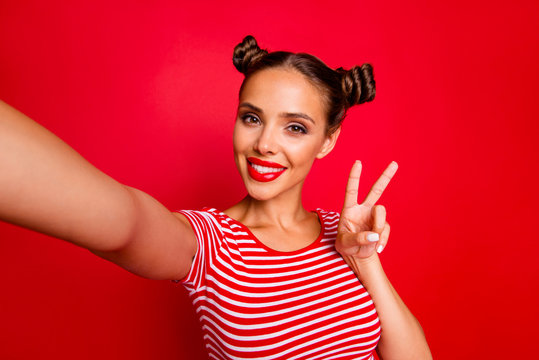Young woman with funky mood making v-sign and selfie on the front camera of smartphone, playful and happy isolated on red background