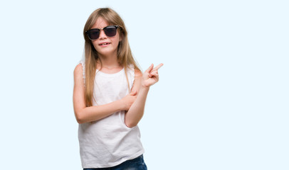 Young blonde toddler wearing sunglasses very happy pointing with hand and finger to the side