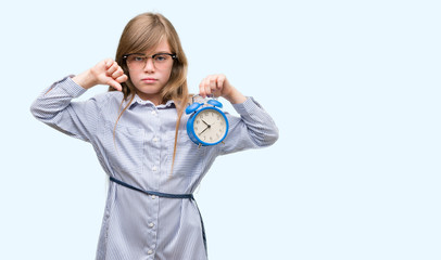 Fototapeta premium Young blonde child holding alarm clock with angry face, negative sign showing dislike with thumbs down, rejection concept