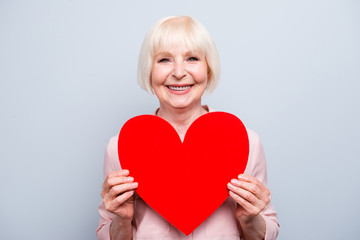 Portrait of old adult blonde caucasian glad lady holding hands big red paper heart shape, smiling...