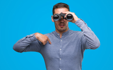Fototapeta premium Handsome young man looking through binoculars with surprise face pointing finger to himself