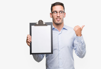 Handsome young business man showing a clipboard pointing with hand and finger up with happy face...