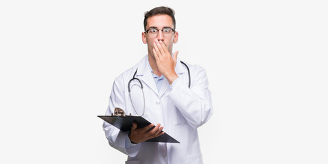 Handsome young doctor man holding a clipboard cover mouth with hand shocked with shame for mistake,...