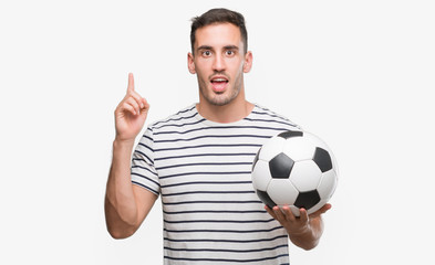 Handsome young man holding soccer football surprised with an idea or question pointing finger with...