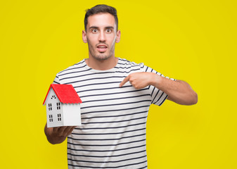 Handsome real estate agent holding a house with surprise face pointing finger to himself
