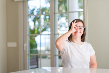 Down syndrome woman at home with happy face smiling doing ok sign with hand on eye looking through fingers