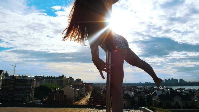 Long-haired sexy woman doing exercises on the pylon against the cityscape at sunset