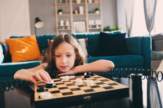 Lifestyle Shot Of Smart Kid Girl Playing Checkers At Home. Board Games For Kids Concept, Candid Series With Real People In Modern Interior