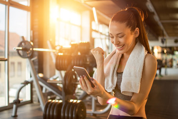 Euphoric young woman with beautiful smile reading good news on mobile phone and celebrating success during workout break in gym