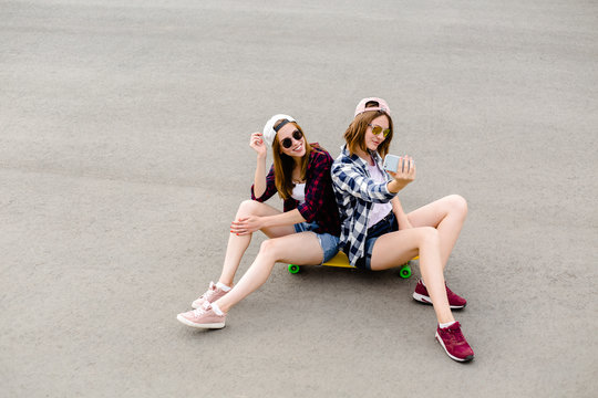 Two young happy girl friends in hipster outfit sitting together on longboard and making selfie on phone