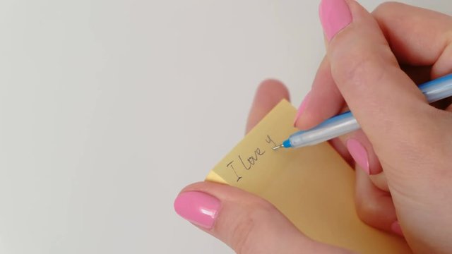Close up woman's hands writing note "I love you" kisses the paper, leaving a trail of lips of lipstick and sticks it on the wall.