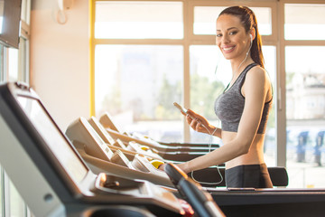 Happy fit woman in fitness wear with phone and earphones on treadmill machine in the gym