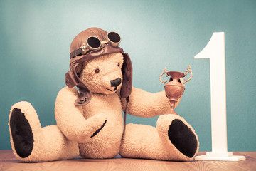 Retro Teddy Bear toy in leather pilot's hat and vintage goggles sitting with award winner's trophy cup and big wooden number one front mint green wall background. Old style filtered photo