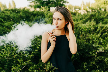 Attractive young woman in casual stylish clothes smoking an electronic cigarette, standing near the green bushes, looking at camera, outdoors.