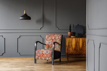 Patterned armchair next to wooden cabinet in grey living room interior with black lamp. Real photo
