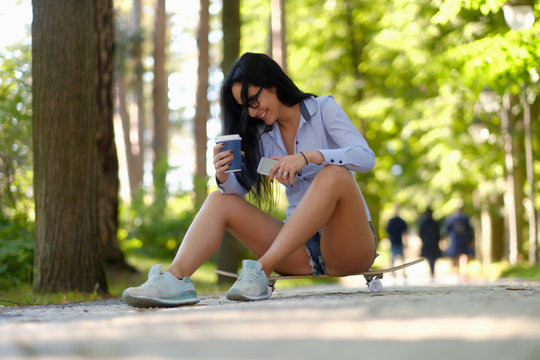 Laughing sexy brunette girl in glasses wearing a shirt and shorts sitting on a skateboard and holds a cup of takeaway coffee and smartphone at the park.